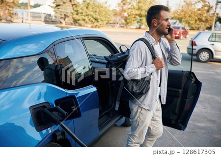 Talking by phone. Man with blue electric car on the charge station Talking by phone. Man with blue electric car on the charge station 128617305