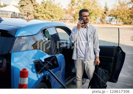 Talking by phone. Man with blue electric car on the charge station Talking by phone. Man with blue electric car on the charge station 128617306