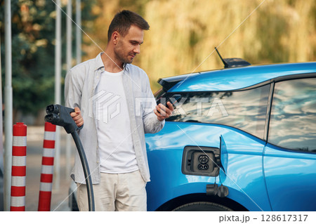 Wire in socket, holding smartphone. Man with blue electric car on the charge station 128617317
