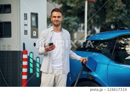 Wire in socket, holding smartphone. Man with blue electric car on the charge station Wire in socket, holding smartphone. Man with blue electric car on the charge station 128617319