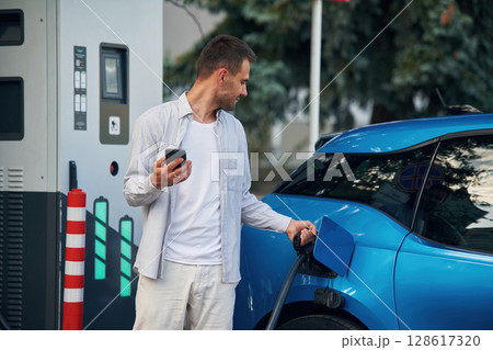 Wire in socket, holding smartphone. Man with blue electric car on the charge station 128617320