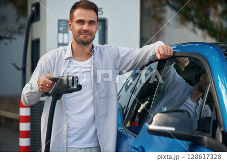 With wire in hand. Man with blue electric car on the charge station With wire in hand. Man with blue electric car on the charge station 128617328