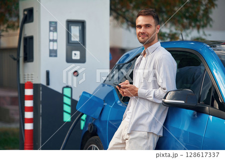 Side view, standing with smartphone, waiting. Man with blue electric car on the charge station 128617337