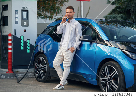 Talking by phone. Man with blue electric car on the charge station 128617343