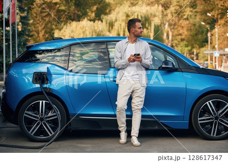 Blue vehicle, holding smartphone. Man with blue electric car on the charge station Blue vehicle, holding smartphone. Man with blue electric car on the charge station 128617347