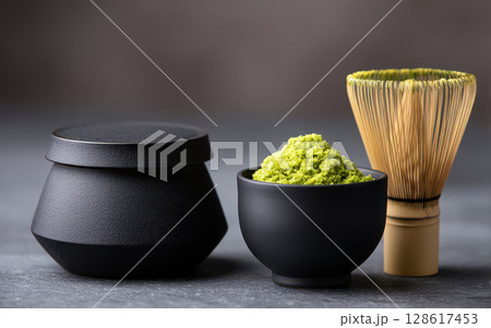 Matcha green tea powder in black bowl with bamboo whisk and container for morning mindfulness routine preparation in soft light 128617453