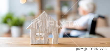Wooden house model on table with blurred elderly person in wheelchair in background symbolizing rehabilitation and retirement home care 128617729