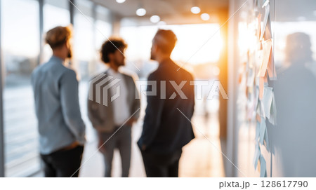 Inclusive workplace culture is shown by three colleagues discussing ideas near wall with sticky notes in modern office with warm sunlight 128617790