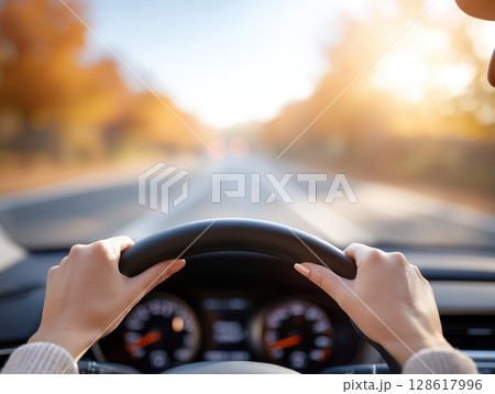 Close up view of hands gripping steering wheel while driving on sunlit road with autumn trees blurred in background, creating warm and calm atmosphere 128617996