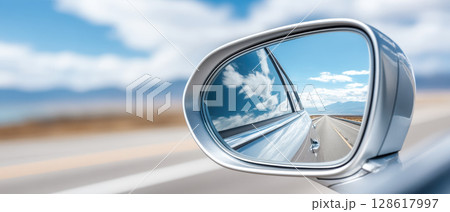 Closeup of car side mirror reflecting clear blue sky with clouds and open road stretching into distance, evoking sense of freedom and travel 128617997