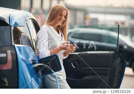 Smartphone in hands. Woman on the electric cars charge station at daytime Smartphone in hands. Woman on the electric cars charge station at daytime 128618120