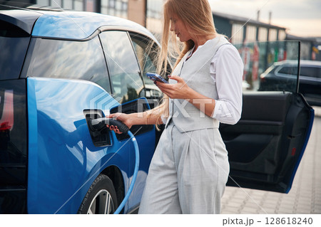 Wire is in the socket. Woman on the electric cars charge station at daytime Wire is in the socket. Woman on the electric cars charge station at daytime 128618240