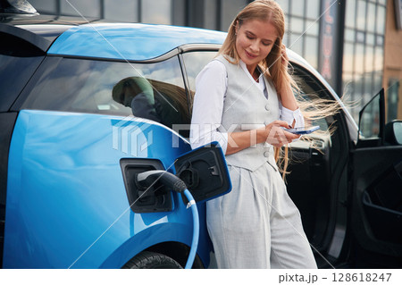 Standing and waiting. Woman on the electric cars charge station at daytime Standing and waiting. Woman on the electric cars charge station at daytime 128618247