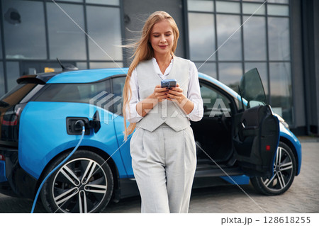 Using smartphone. Woman on the electric cars charge station at daytime Using smartphone. Woman on the electric cars charge station at daytime 128618255