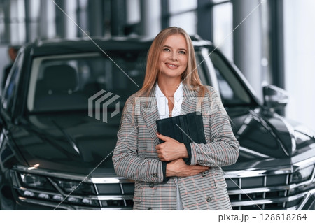 Black colored document in hands. Woman in formal clothes is in the car dealership Black colored document in hands. Woman in formal clothes is in the car dealership 128618264