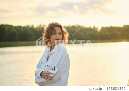 Cute woman with curly hair stands by the lake at sunset Cute woman with curly hair stands by the lake at sunset 128621562