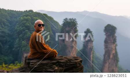 Poster with monk in sunglasses sitting in lotus position or padmasana rock on edge cliff with beautiful mountain view, smiling looking camera, copy space freedom retreat concept away everyday bustle 128623051