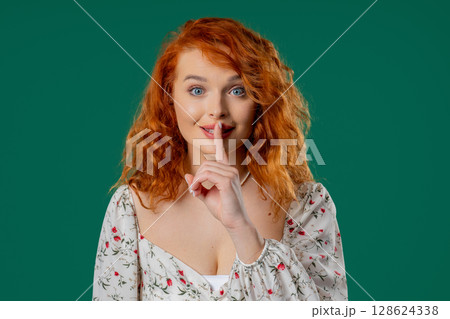 Beautiful woman with natural red curls makes shush shh gesture with finger on lips, silence signaling 128624338
