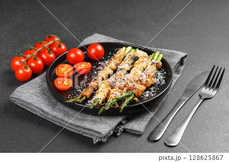 Grilled asparagus wrapped with bacon on black plate with cherry tomatoes on black background 128625867