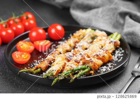 Grilled asparagus wrapped with bacon on black plate with cherry tomatoes on black background Grilled asparagus wrapped with bacon on black plate with cherry tomatoes on black background 128625869
