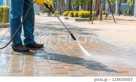 Worker pressure washing a brick surface. Person wearing yellow gloves using a high-pressure cleaner. Cleaning outdoor brick flooring for maintenance. 128630083