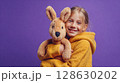 Girl with stuffed animal, smiling. Young child hugging a plush kangaroo on a solid purple backdrop.  Warm and endearing childhood moment. 128630202