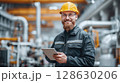Smiling Engineer in Hardhat Holding Tablet. Industry expert oversees operations with tech in a factory, ensuring efficiency and safety with a confident smile. 128630206