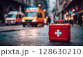 Emergency Medical Response: Close-up of a red first aid kit stands out against the blurred backdrop of an urban street with emergency vehicles in action. 128630210