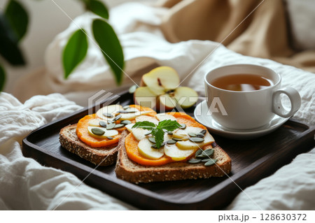 Cozy breakfast is served on a wooden tray featuring creative toasts topped with sweet potato and garnished with apple slices and mint. A cup of tea completes the warm setting 128630372