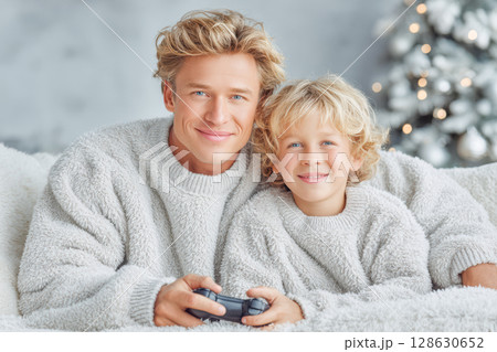 Father and son enjoying video game together in cozy sweaters on a soft couch with festive decor 128630652