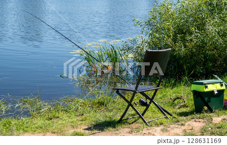 Fishing rod on holder by river with chair and tackle box on green grass 128631169