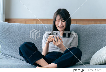 ソファに座ってスマートフォンを使う笑顔の女性 ソファに座ってスマートフォンを使う笑顔の女性 128631750