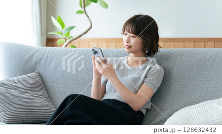 ソファに座ってスマートフォンを使う笑顔の女性 ソファに座ってスマートフォンを使う笑顔の女性 128632432