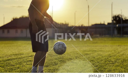 Male feet of professional footballer kicking ball at green field. Legs of young man juggling soccer ball on stadium at sunset. Sportsman practicing tricks outdoor. Freestyle football concept 128633916