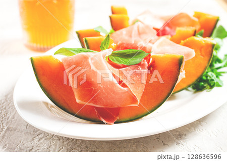 Cantaloupe melon with prosciutto and fresh basil, on a white plate, natural light, 128636596