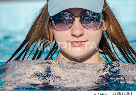 Teenage girl wearing sunglasses and a cap relaxing in a swimming pool, with wet hair and calm expression, enjoying a sunny summer day 128638361