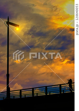 橋の上に立つ街灯と夕焼けを撮る女性 橋の上に立つ街灯と夕焼けを撮る女性 128640053