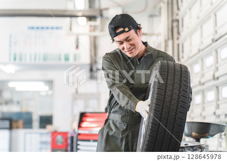 自動車整備工場でタイヤを運ぶ男性メカニック（肉体労働・重労働・重い・腰痛） 128645978