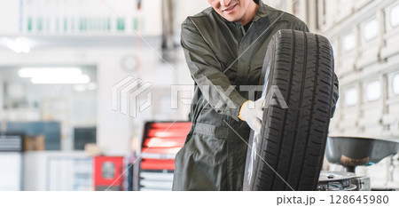 自動車整備工場でタイヤを運ぶ男性メカニック（肉体労働・重労働・重い・腰痛） 128645980