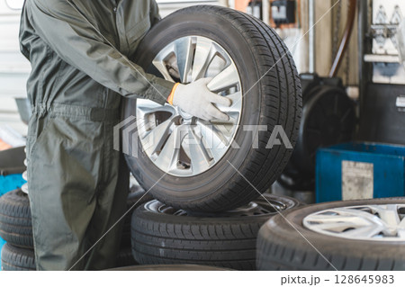 自動車整備工場でタイヤを運ぶ男性メカニック（肉体労働・重労働・重い・腰痛） 128645983