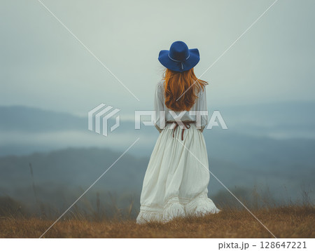 Elegant woman, seen from behind, gazing at misty mountains. She seems reflective and thoughtful, taking in serene landscape in her white dress and blue hat Elegant woman, seen from behind, gazing at misty mountains. She seems reflective and thoughtful, taking in serene landscape in her white dress and blue hat 128647221