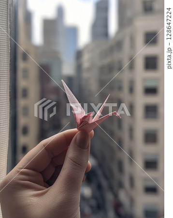 Delicate pink origami crane is held up to window with skyscrapers in background, evoking sense of peace and hope against urban landscape Delicate pink origami crane is held up to window with skyscrapers in background, evoking sense of peace and hope against urban landscape 128647224