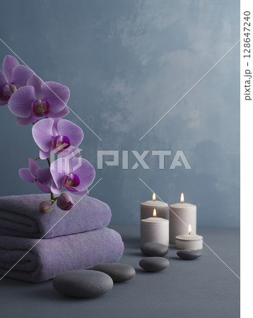 Scene shows stack of purple orchid flowers atop soft towels, lit by candles, creating soothing and peaceful ambiance for relaxation 128647240
