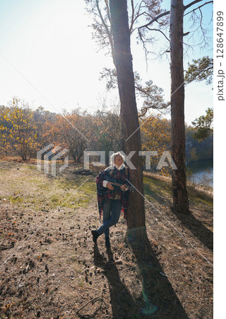 Beautiful blonde girl playing guitar ukulele and singing relaxing enjoying outdoors on sunny autumn day. Hippie woman in boho style training playing musical instrument. Fall leaves 128647899