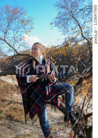 Portrait of smiling caucasian girl plays ukulele in autumnal park. Young woman plays guitar musical instrument outside in nature fall time. Audio music healing 128647900