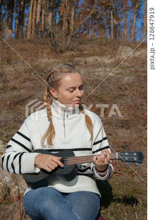 Blonde woman playing guitar ukulele outdoors in autumn forest. Concept of sound therapy, mental health and wellness rituals. Calmness tranquility audio-sensory practices. Aura farming Blonde woman playing guitar ukulele outdoors in autumn forest. Concept of sound therapy, mental health and wellness rituals. Calmness tranquility audio-sensory practices. Aura farming 128647919