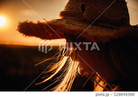 Close-up of a teen girl in a straw hat at sunset, warm light highlighting her hair and profile in a peaceful countryside moment 128648360