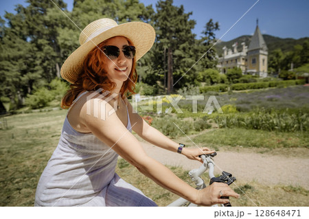 A woman wearing a straw hat and sunglasses is riding a bicycle in a park A woman wearing a straw hat and sunglasses is riding a bicycle in a park 128648471