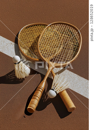 Two wooden badminton rackets with yellow strings and two shuttlecocks on tennis court with white line, sunlight casting shadows, creating warm and sporty atmosphere 128652019