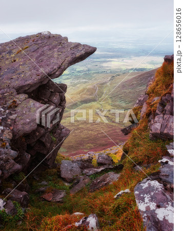 Viewing lush valley in Ireland's Coomloughra Horseshoe through rocky outcrop on a cloudy day, green fields and blue sky in background. Viewing lush valley in Ireland's Coomloughra Horseshoe through rocky outcrop on a cloudy day, green fields and blue sky in background. 128656016
